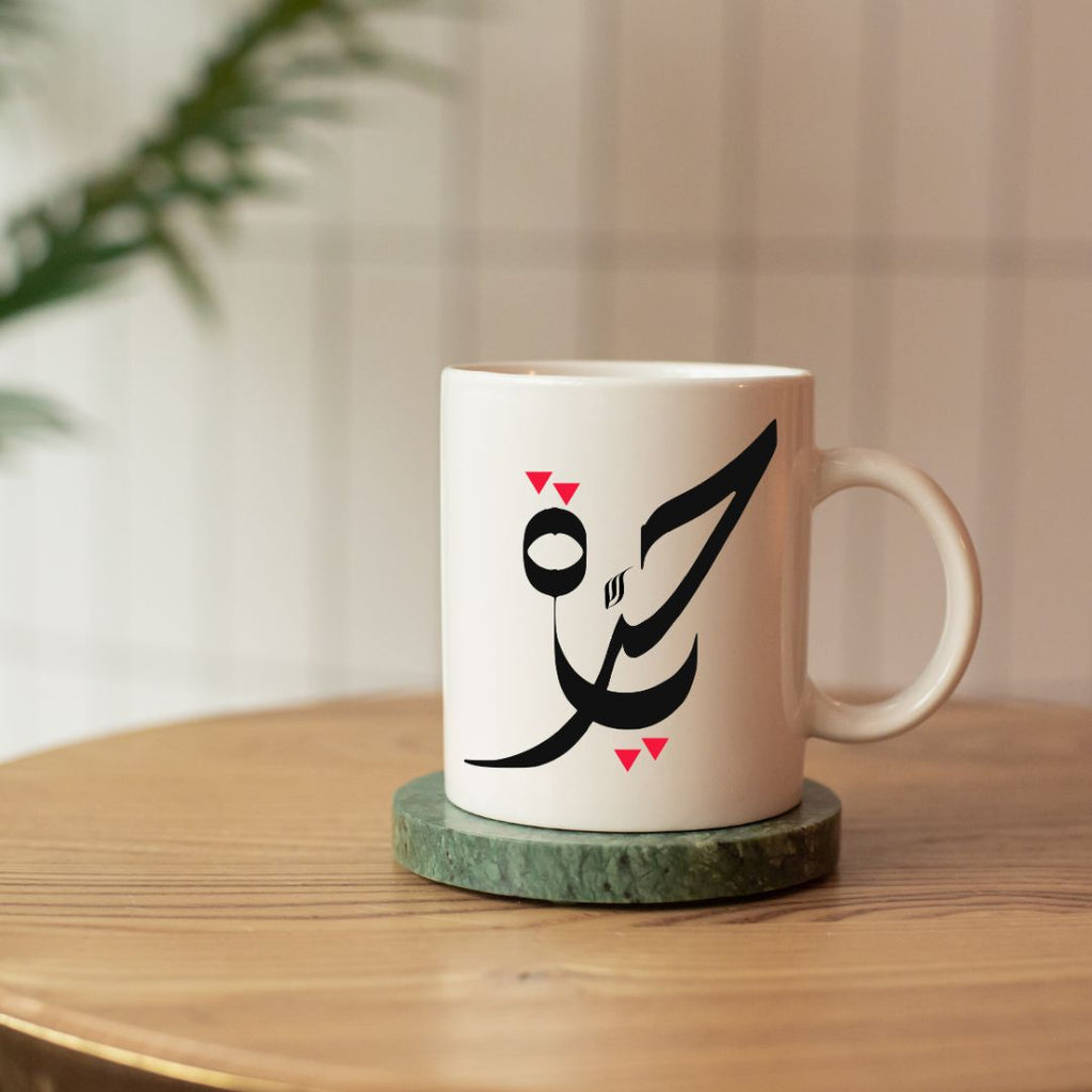 White mug with black Arabic calligraphy Hurriyah and red triangles on a wooden table.
