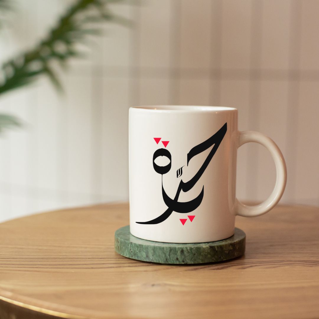 White mug with black Arabic calligraphy Hurriyah and red triangles on a wooden table.